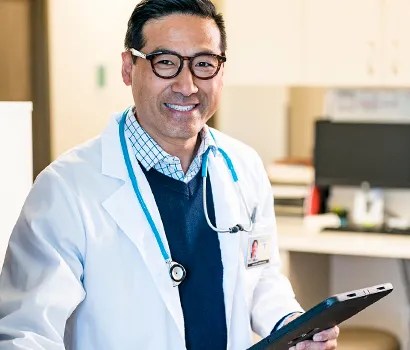 Photo of a smiling doctor wearing glasses, a white lab coat, stethoscope, and holding a tablet in a medical office setting